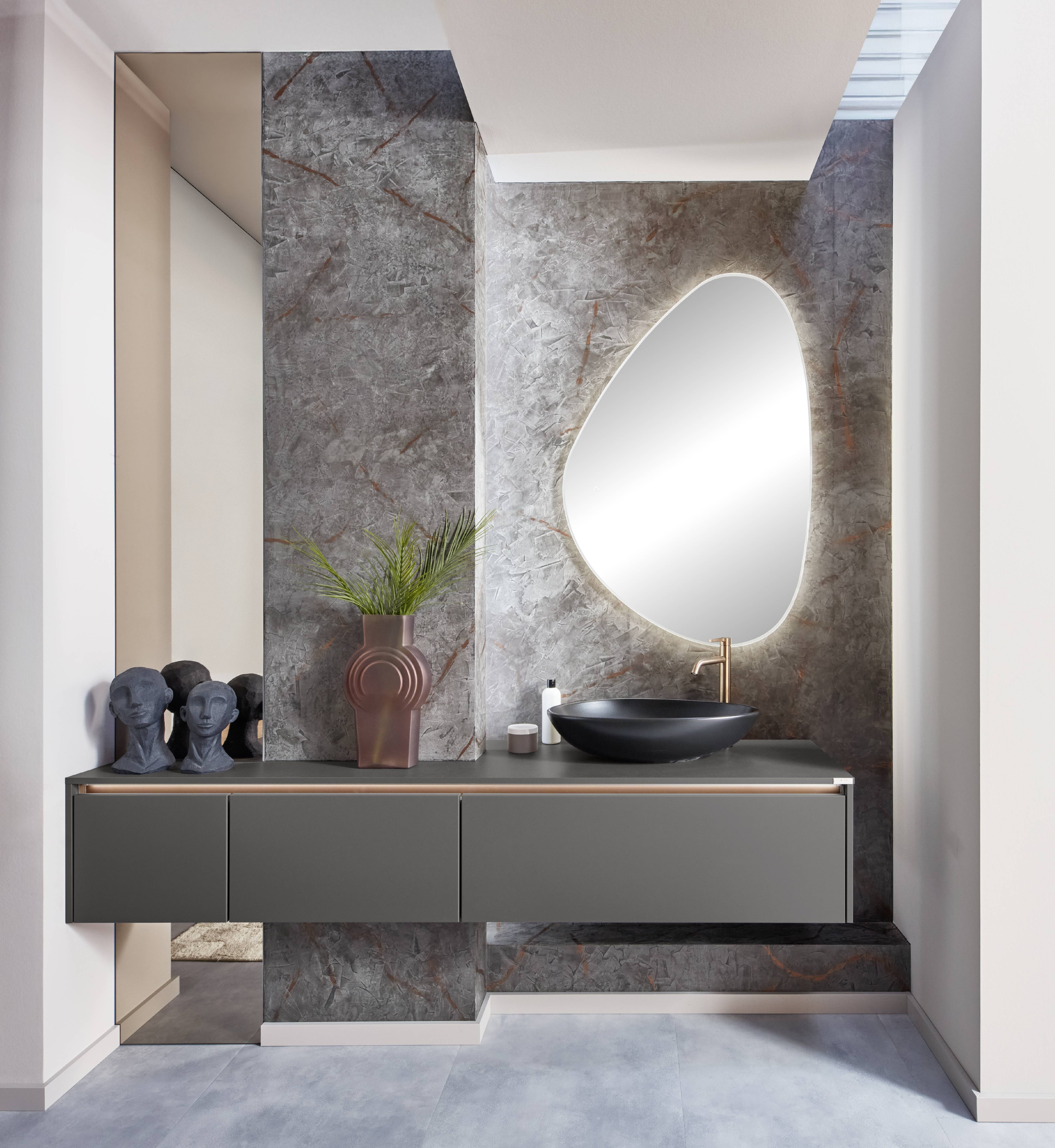 Modern Floating Vanity with Stone-Effect Wall and Statement Mirror Modern bathroom with a floating charcoal vanity, black vessel sink, warm metallic faucet, textured stone-effect wall panels, and an organic-shaped backlit mirror.