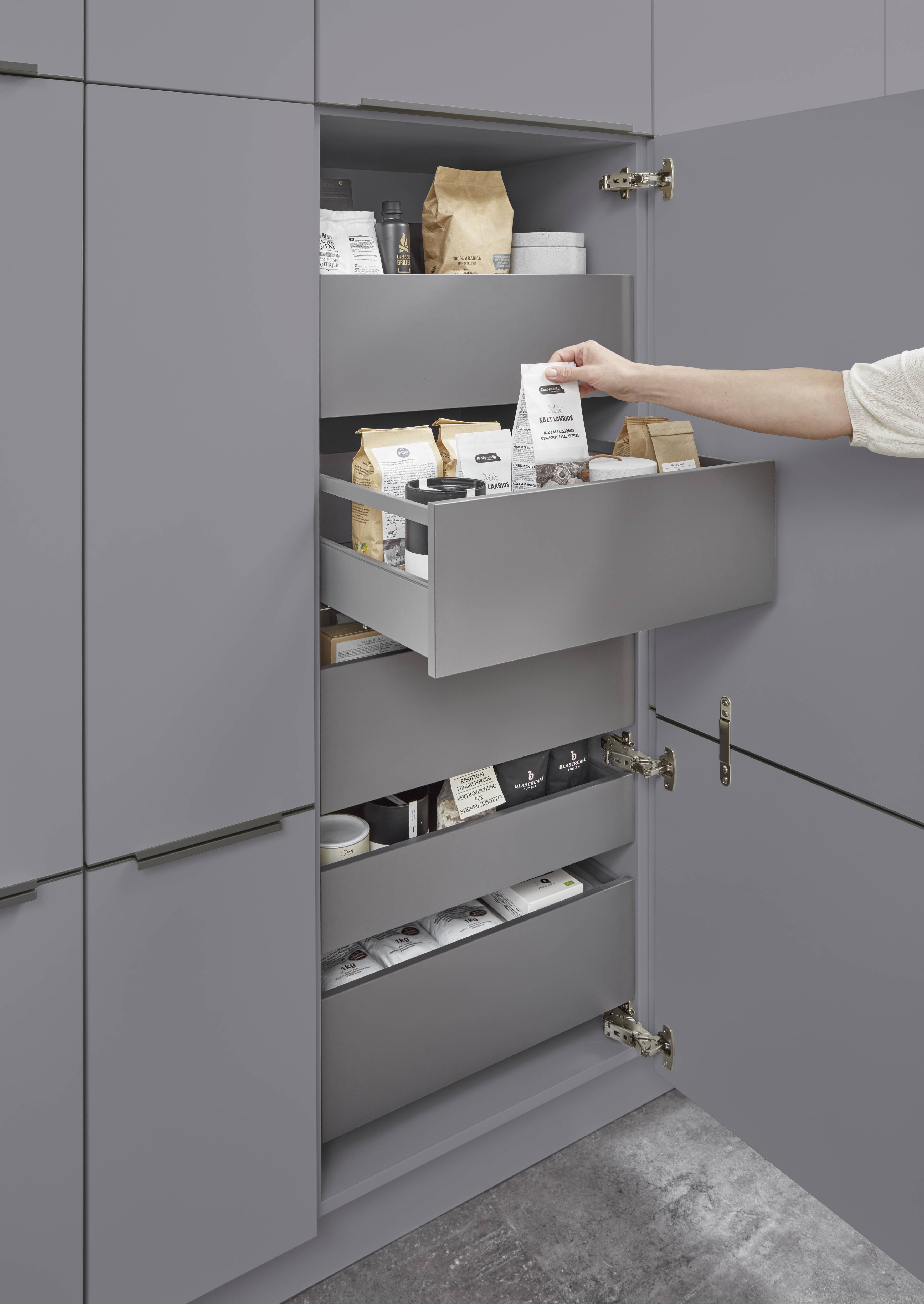 Tall pantry cabinet with pull-out drawers in a modern grey kitchen, with a hand reaching for a pantry item