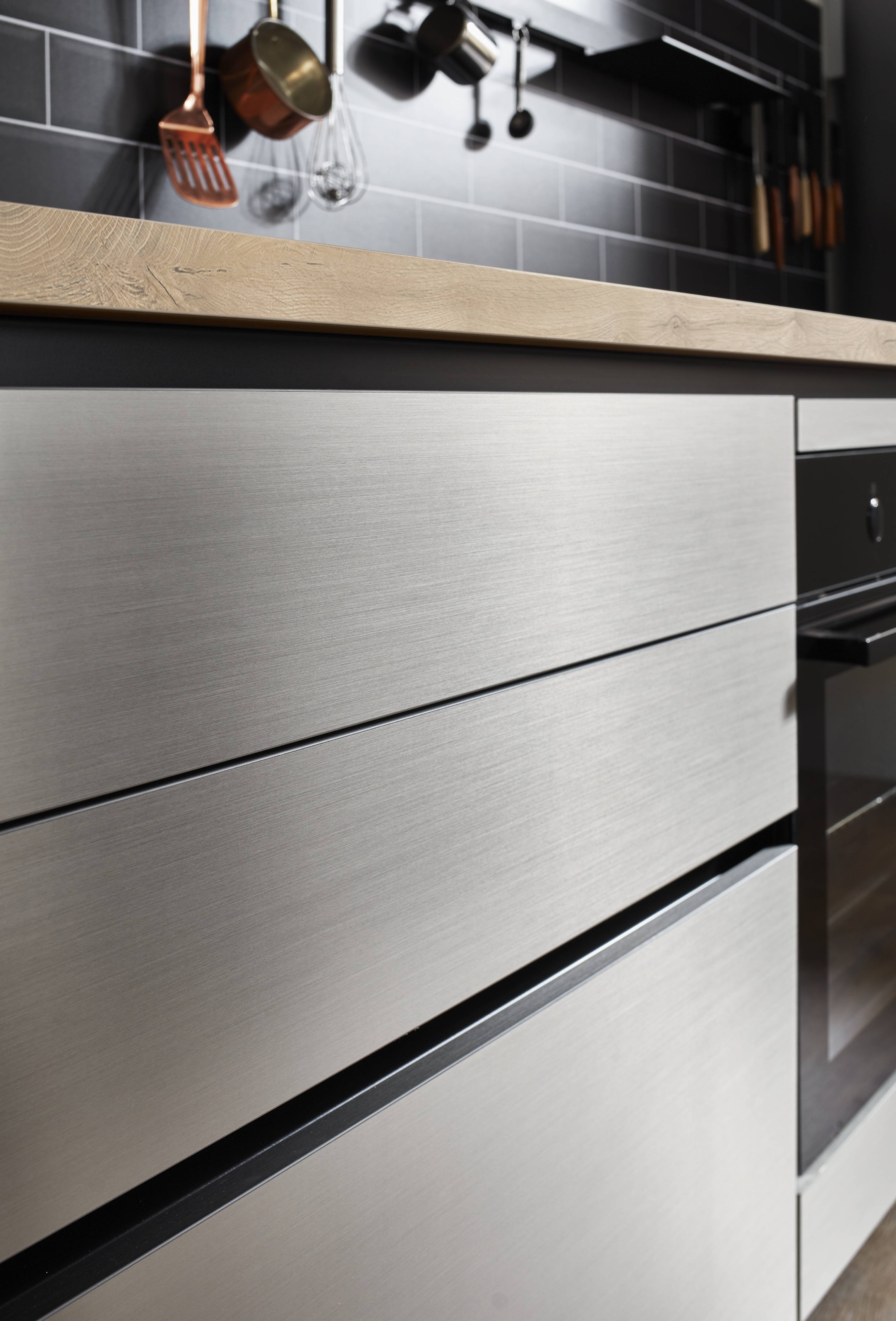 Close-up of brushed metal kitchen drawer fronts beneath a warm wood countertop, with hanging utensils and a tiled backsplash in the background.