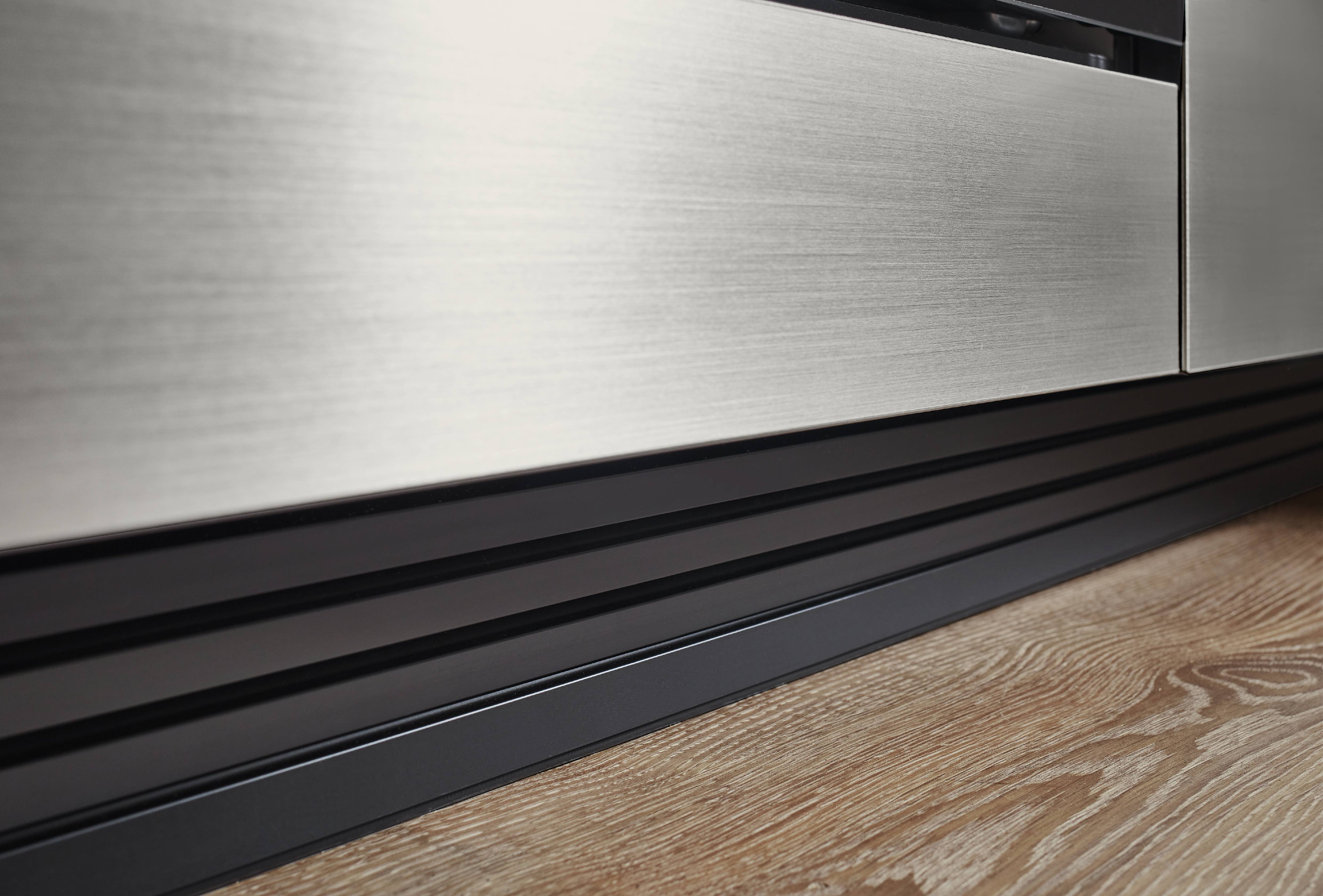 Close-up of a brushed metal cabinet front with a black grooved toe-kick, adding a sleek industrial detail over wood-look flooring.