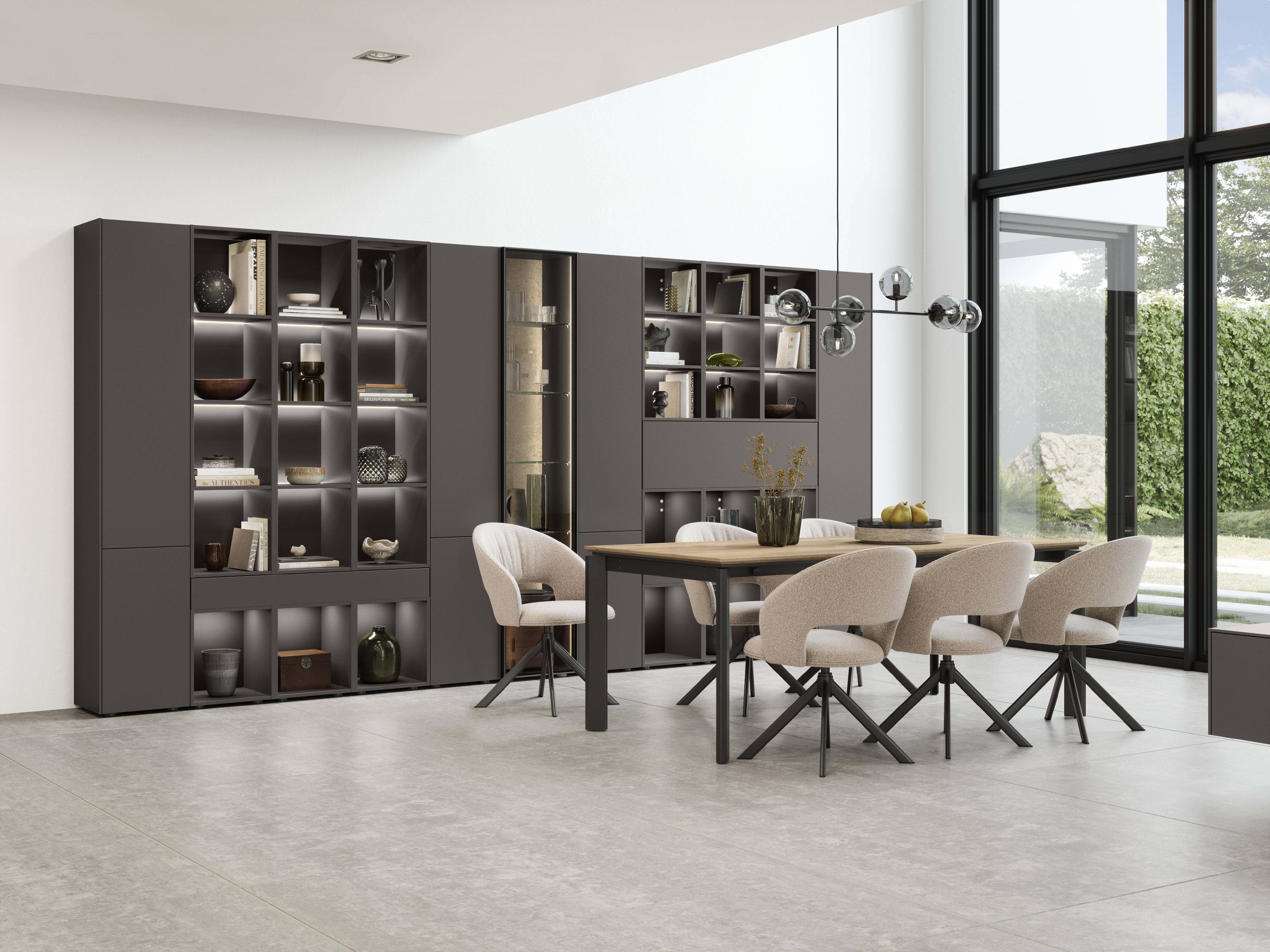 A modern dining room featuring a wooden table with six upholstered chairs, a wall of built-in shelving with decorative items and soft lighting, and large floor-to-ceiling windows that bring in natural light.