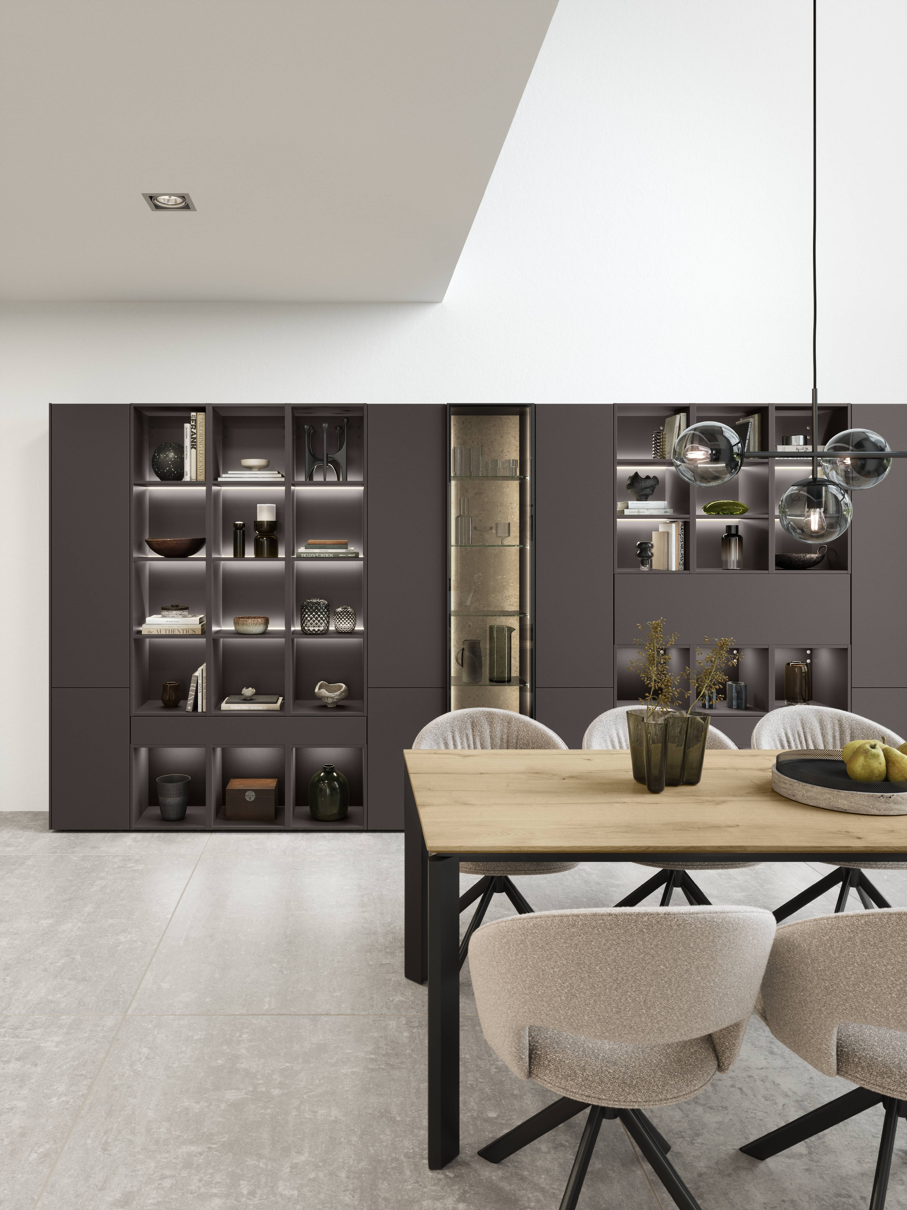 A modern dining space featuring sleek, minimalist design elements with warm neutral tones. The room showcases a custom built-in shelving unit in a deep taupe finish, styled with decorative accents, books, and soft integrated lighting. A natural wood dining table with black metal legs is paired with upholstered swivel chairs in a soft beige fabric, creating a clean yet inviting atmosphere. Contemporary pendant lighting with glass globe details adds an elegant touch, while the open layout and high ceilings enhance the airy, sophisticated feel of the space.