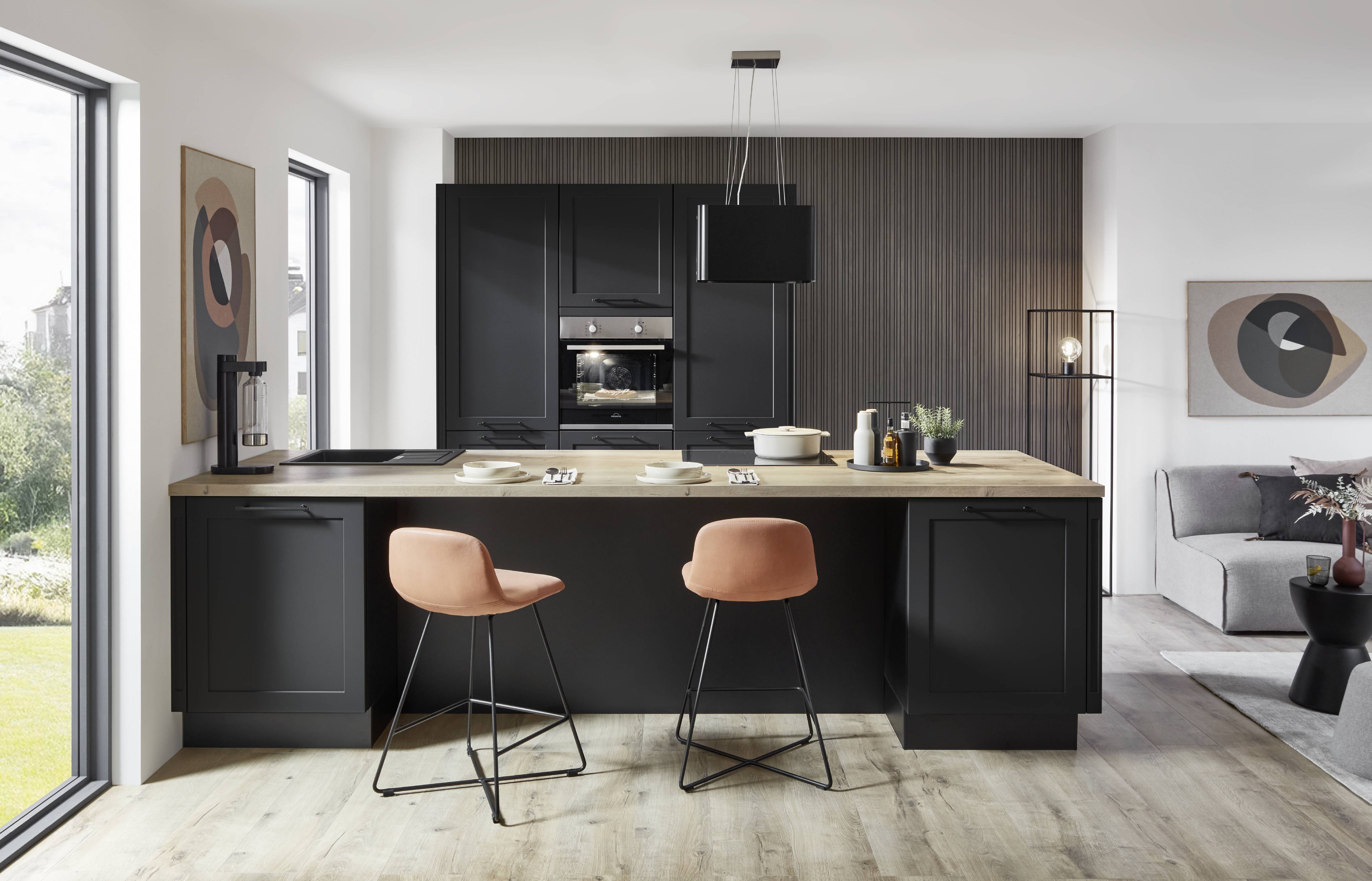 Modern German kitchen featuring matte black cabinetry, a spacious wood-topped island with seating, integrated appliances, pendant lighting, and an open-concept living area.
