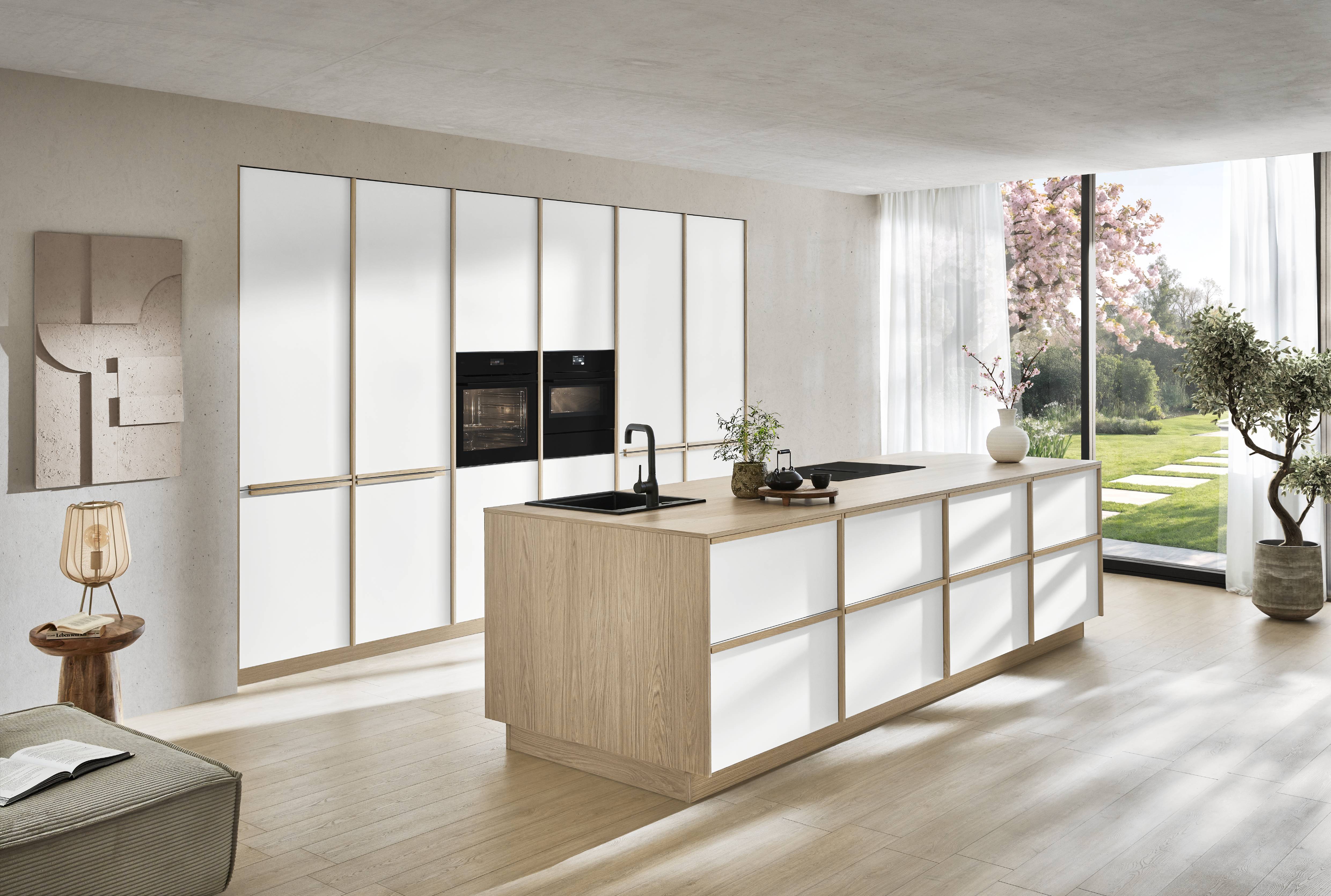 noblessa USA - Modern German Kitchen with White Cabinetry and Wood Accents Modern German kitchen with white cabinetry, warm wood accents, a spacious island, integrated appliances, and large windows overlooking a landscaped garden.
