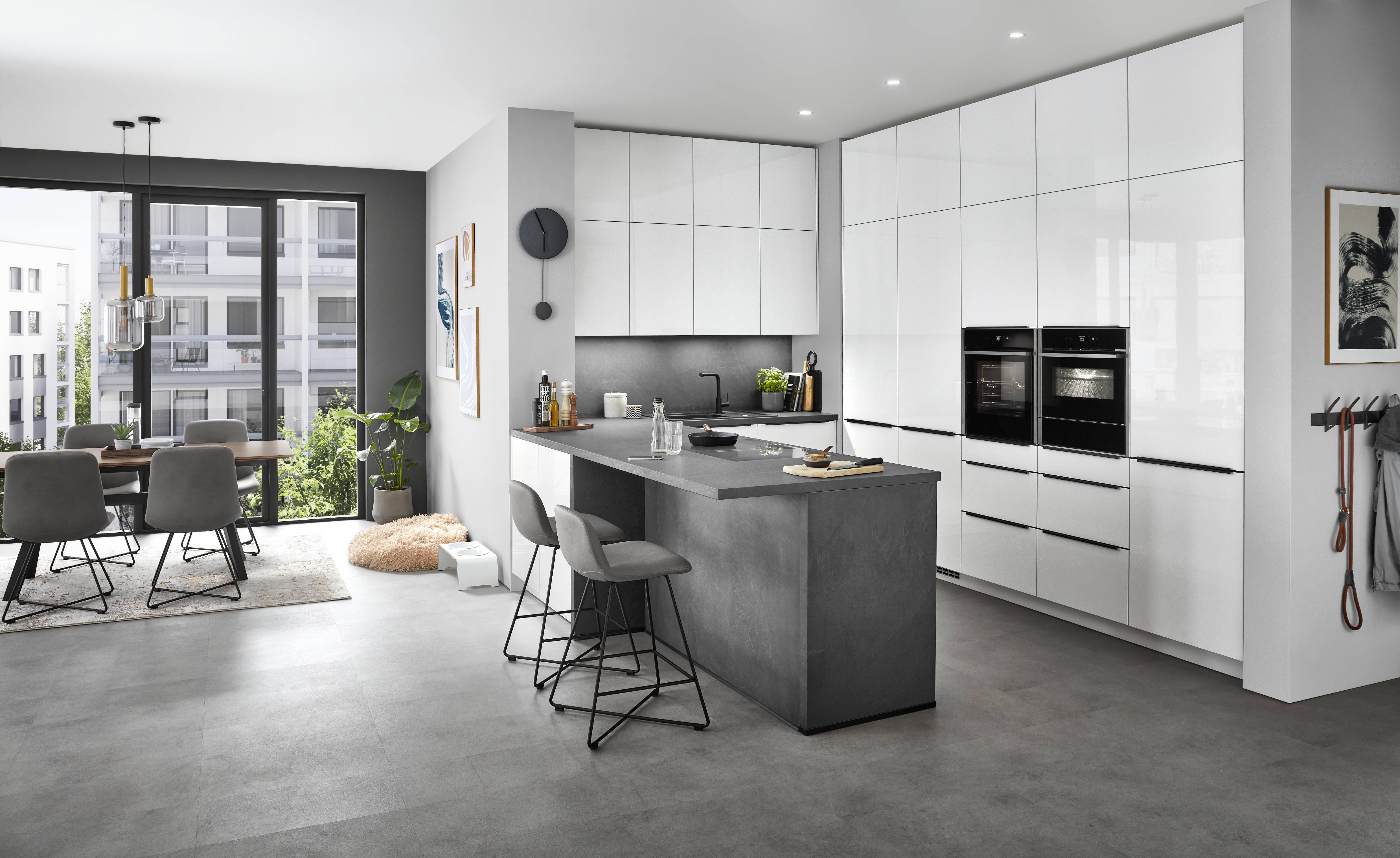 Modern Noblessa USA German kitchen with sleek white cabinetry, dark gray island, integrated double ovens, minimalist finishes, and an open dining area with large windows.