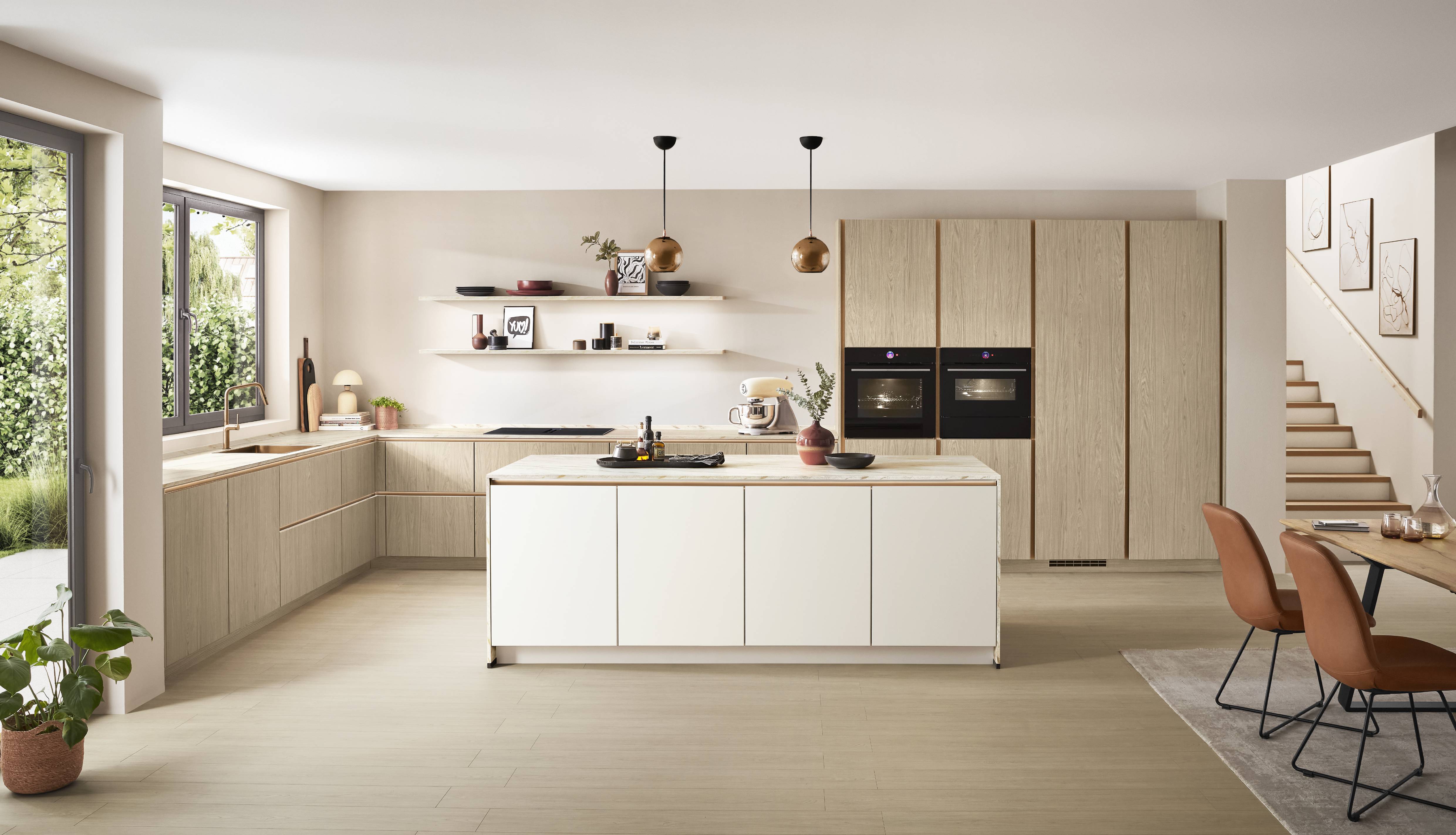 Modern German kitchen with light oak cabinetry, a white island, integrated double ovens, open floating shelves, and large windows that create a bright, airy, open-concept living space.
