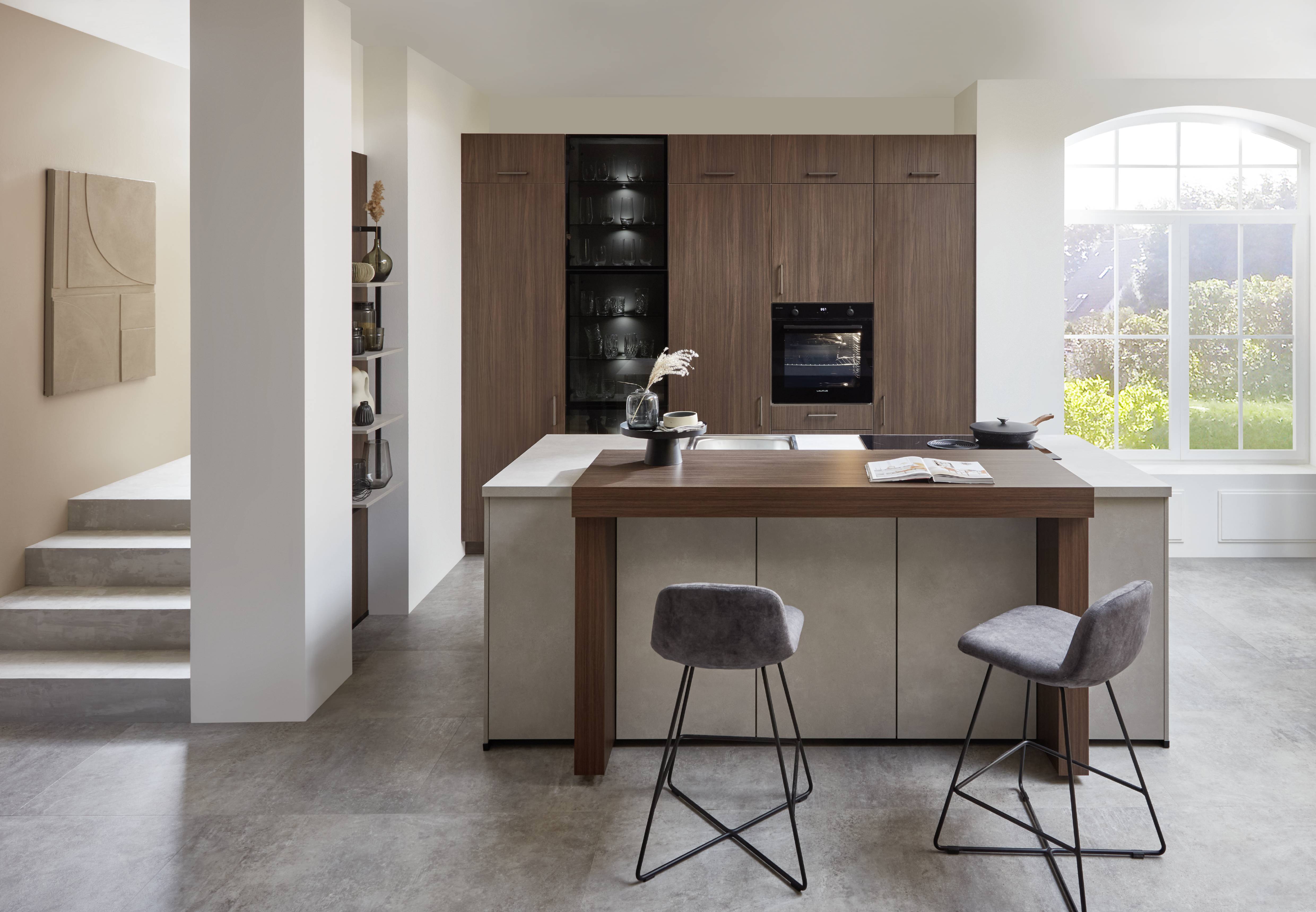 Modern German kitchen with rich wood cabinetry, an integrated oven, glass-front storage, a spacious island, and seating overlooking a large window.