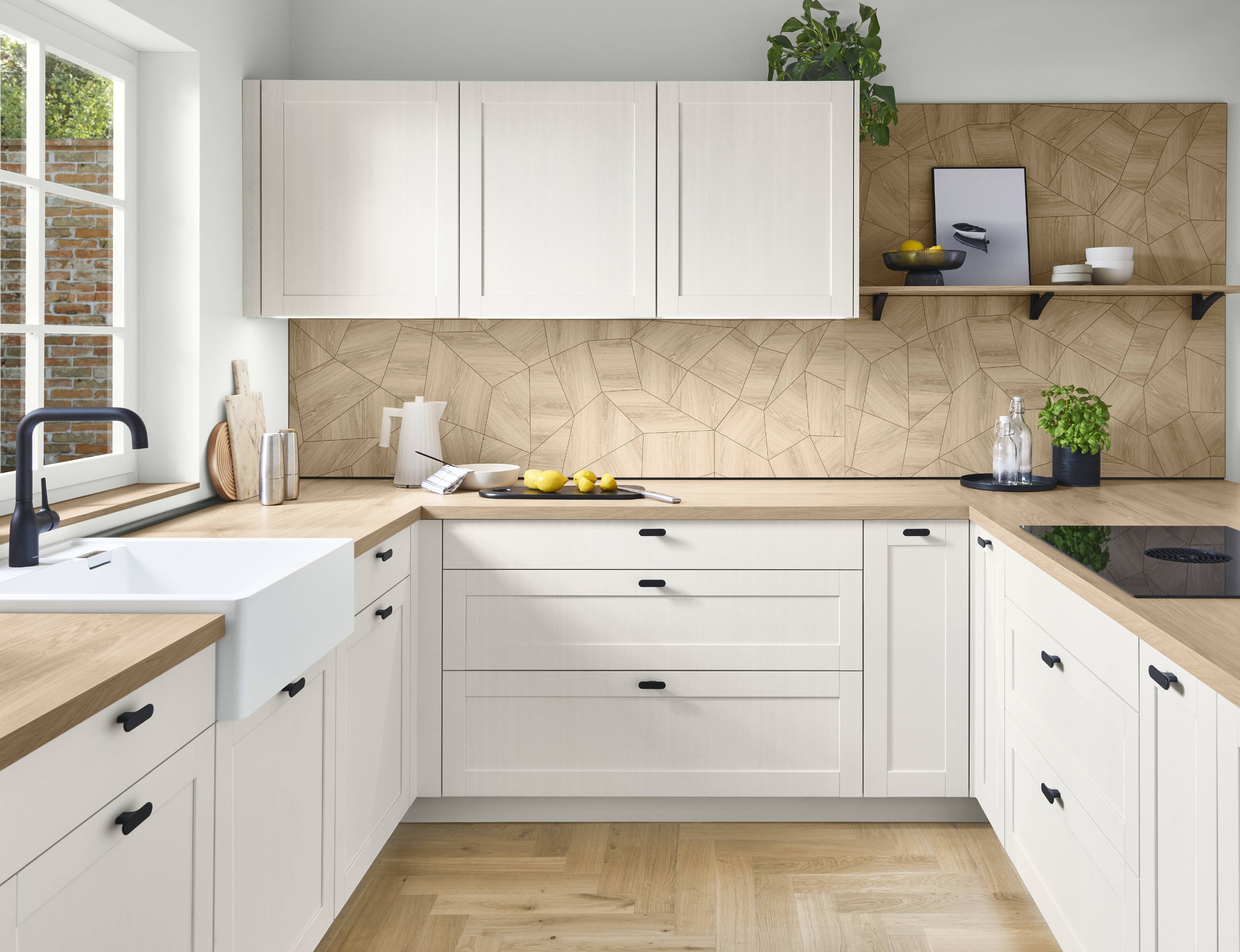 Bright modern German kitchen featuring white shaker-style cabinets, light wood countertops, a geometric wood backsplash, black cabinet handles, a farmhouse sink, open shelving, and natural wood herringbone flooring. The U-shaped layout creates a clean, contemporary, and functional cooking space.