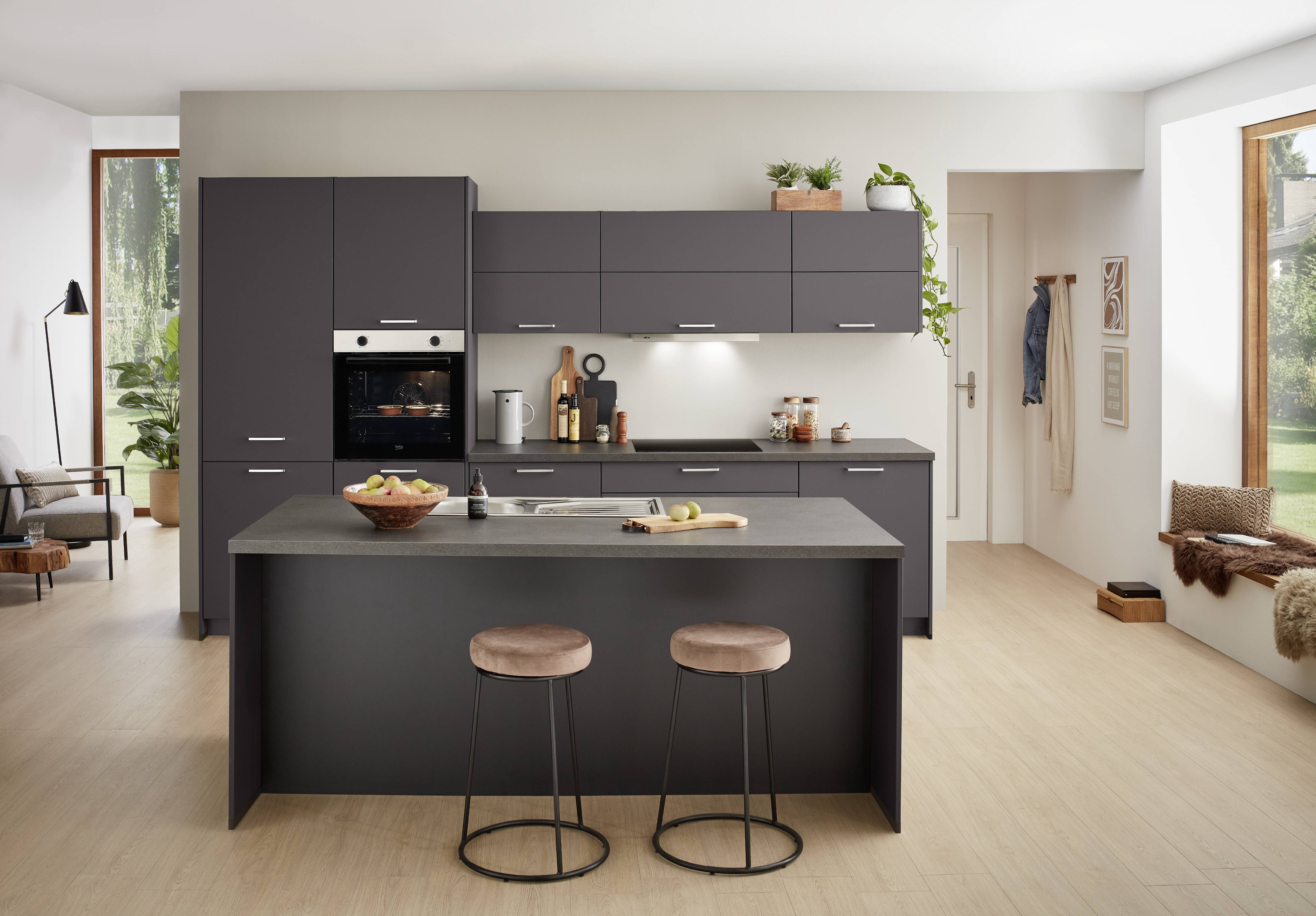 Contemporary kitchen featuring dark gray cabinets, a built-in oven, and a large central island with a gray countertop. Two cushioned bar stools sit at the island, while under-cabinet lighting illuminates the workspace. Decorative plants, wooden accents, and light wood flooring create a warm, modern interior.