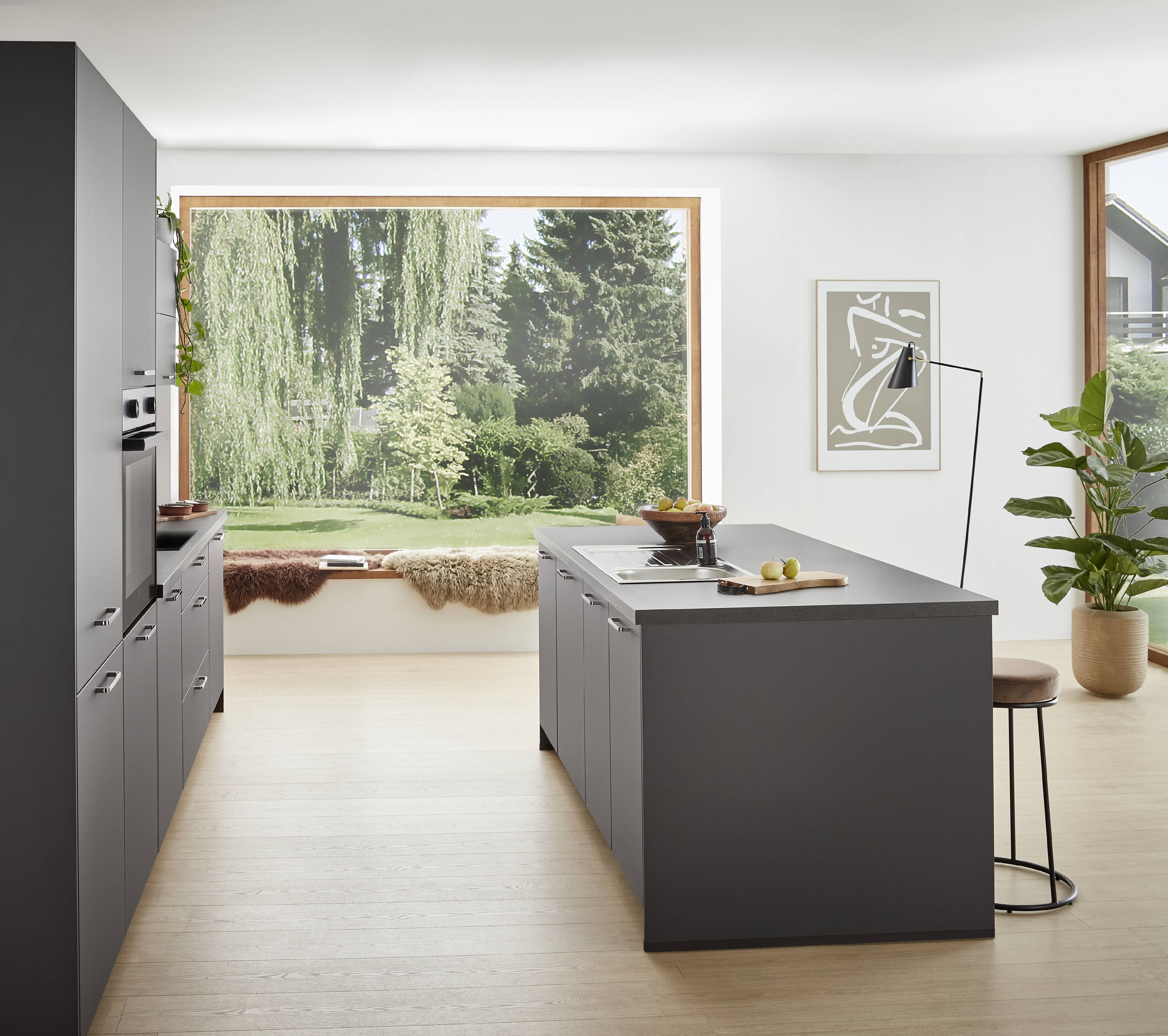 Modern minimalist kitchen featuring matte dark cabinetry, a central island with integrated sink, light wood flooring, and a large picture window overlooking lush green trees and garden landscaping.