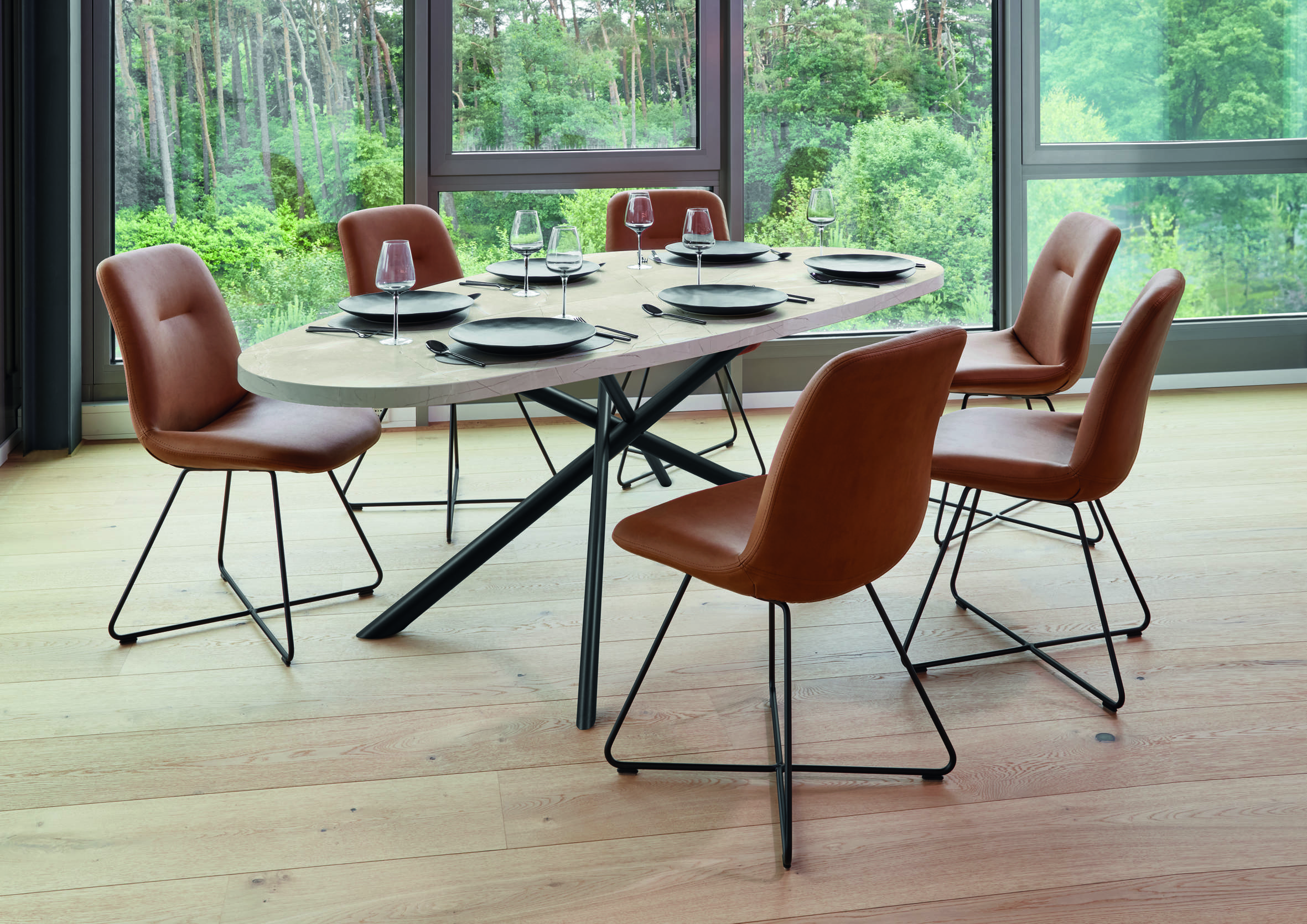 Modern dining room with an oval light-wood table set for six, surrounded by brown leather chairs with black metal legs, positioned beside large floor-to-ceiling windows overlooking a lush forest.